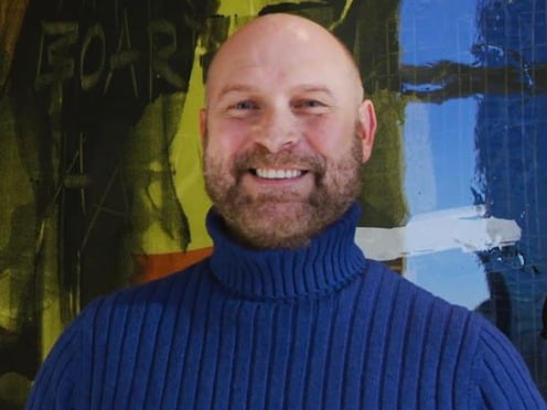 A man wearing a blue turtleneck smiles at the camera.