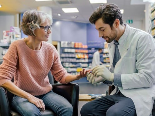 A pharmacist sits in a chair across from a patient and pricks her finger for a blood test