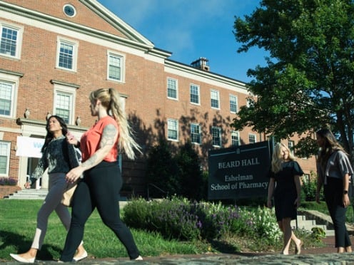 Two women walk down a sidewalk past a pharmacy school.