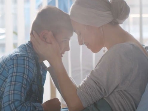 A female cancer patient sits in bed with a scarf around her head and embraces a young boy sitting across from her