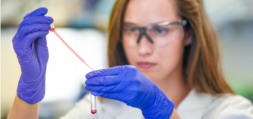 Person in lab coat and blue gloves holding a pipette and test tube with red liquid.