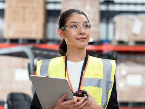 A female warehouse worker wearing safety glasses and a safety vest stands inside a facility holding a clipboard and scanner