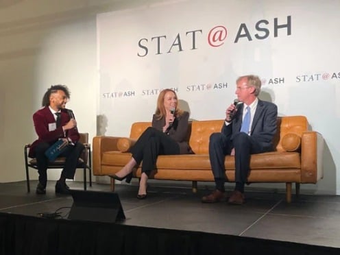Three people sit on a stage discussing under a "STAT@ASH" sign, with a presentation screen nearby.