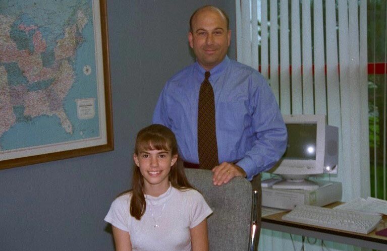 A man in a blue shirt stands behind a girl in a white shirt and checkered skirt in an office.