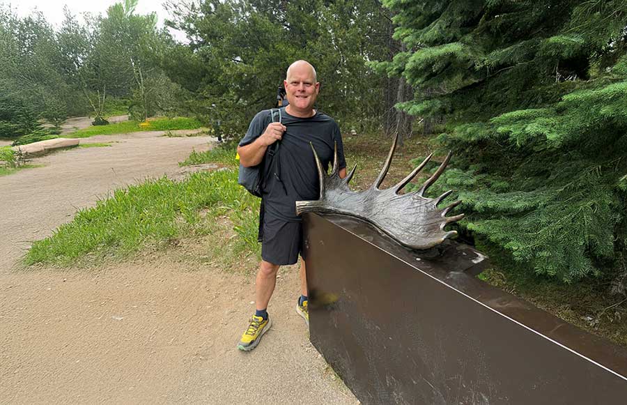 A person stands beside a large moose antler display in a wooded outdoor park.