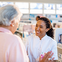 A pharmacist in a white coat assists an elderly customer in a pharmacy.