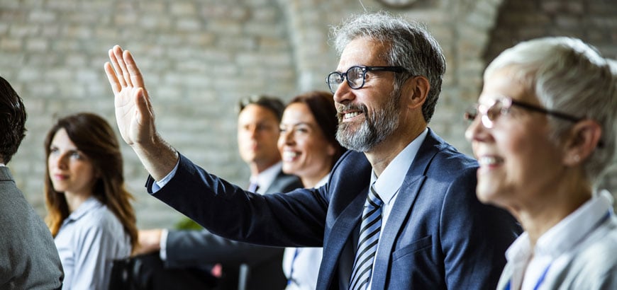 A man in a suit raising his hand in a meeting with other attendees.