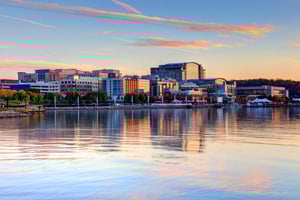 City skyline with buildings reflecting on a calm river at sunset.