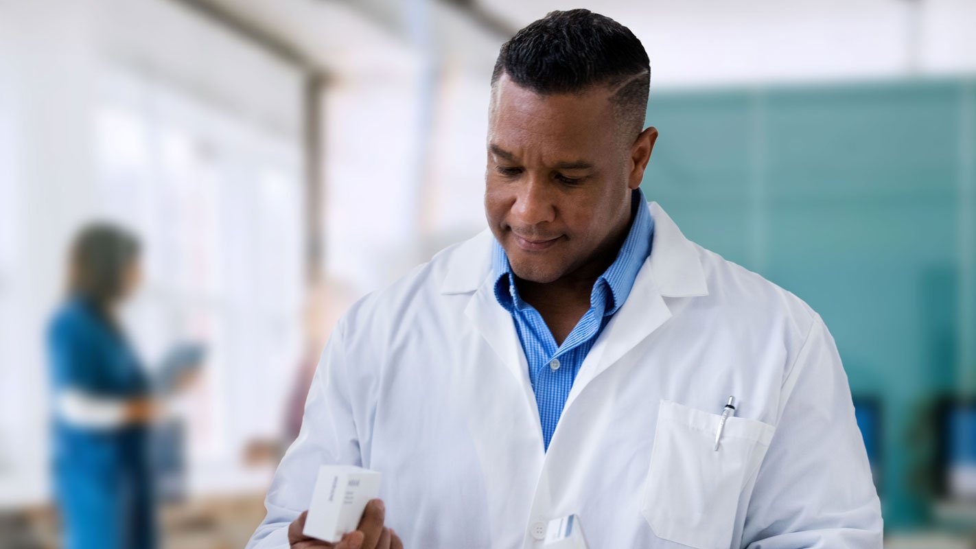 A doctor in a white coat holds a small box, with blurred medical staff in the background.